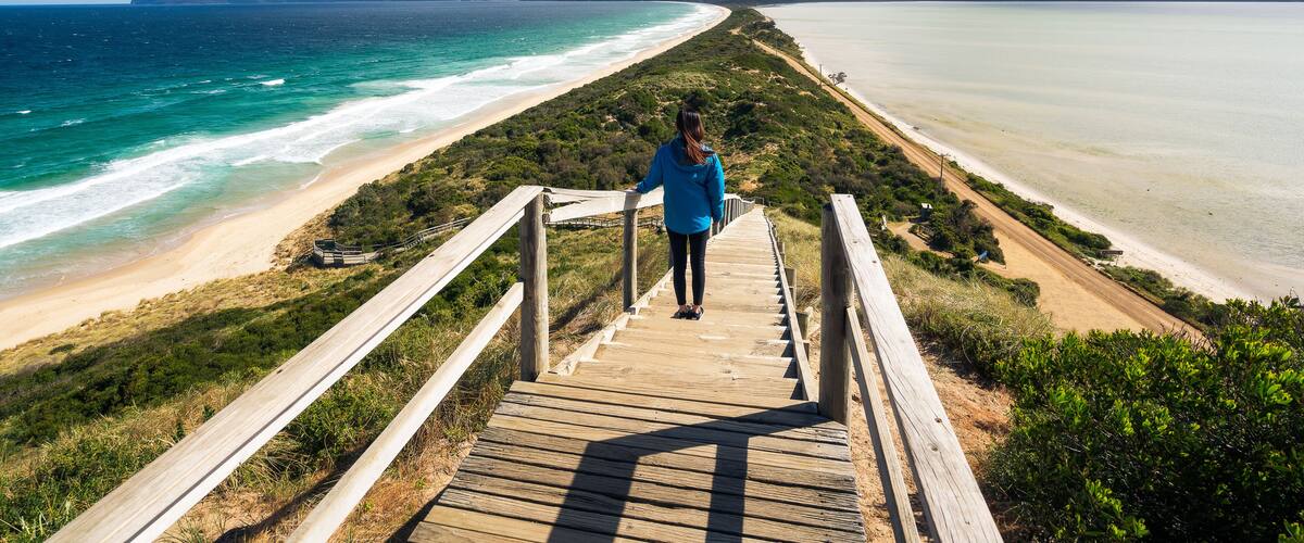 A wooden boardwalk leading to a beach with clear turquoise waters and distant mountains.