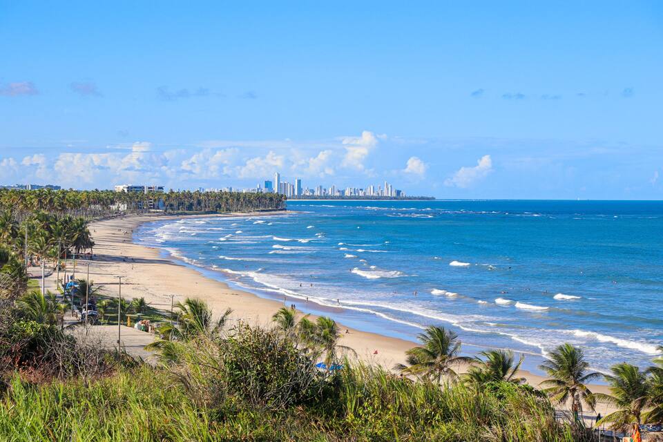 Uma praia com palmeiras, ondas do oceano e um horizonte urbano ao longe.