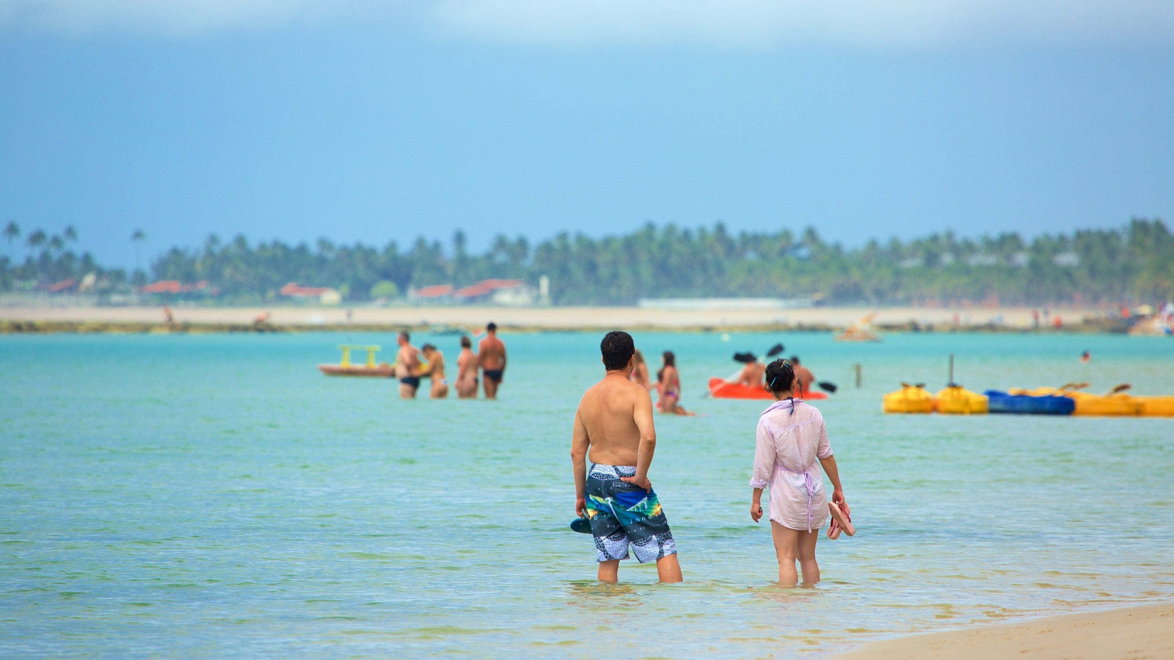 A beach with clear turquoise water, people in swimwear, and a distant shoreline with palm trees.