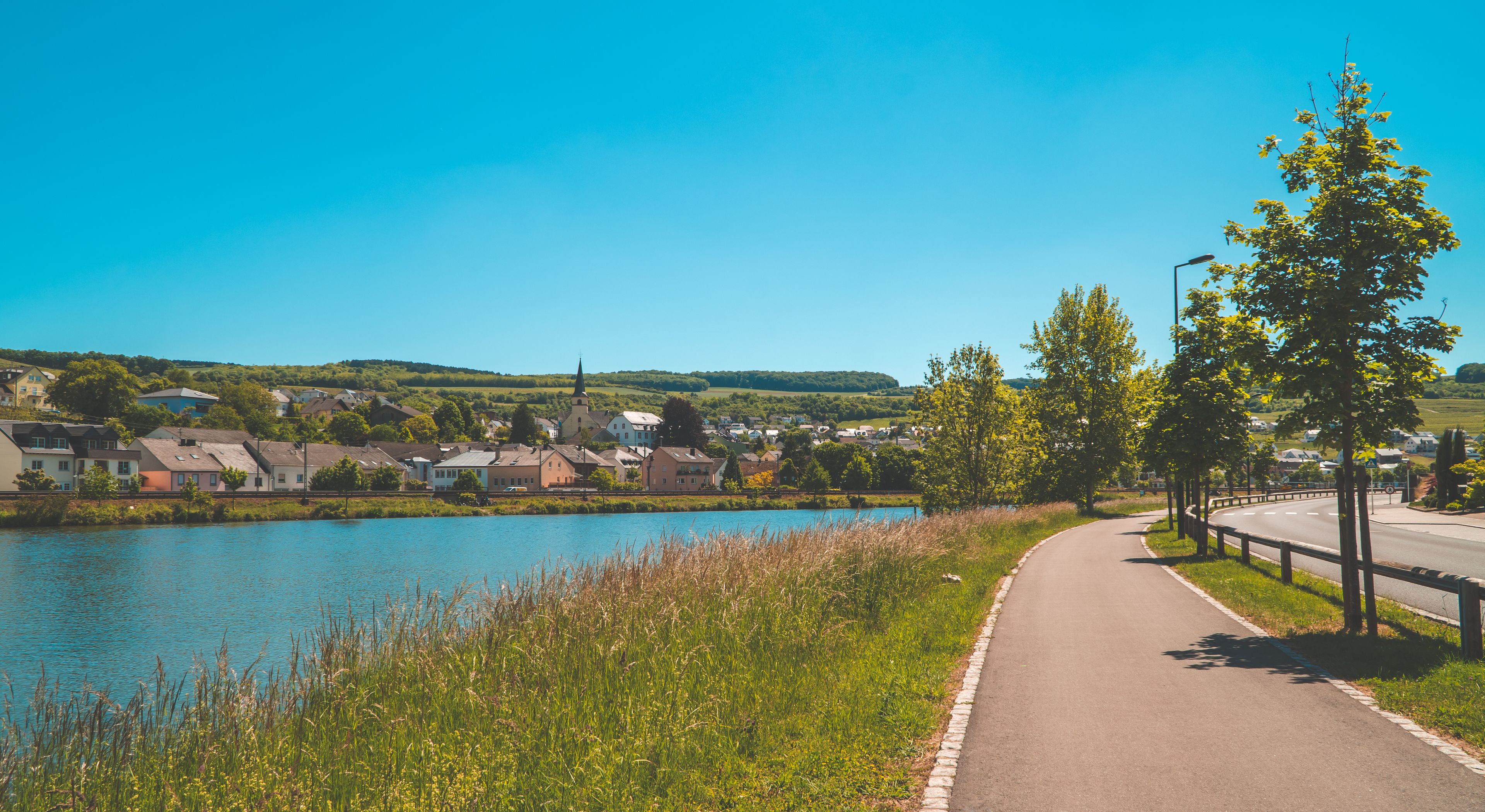 A riverside pathway with houses, a church, and a clear blue sky.