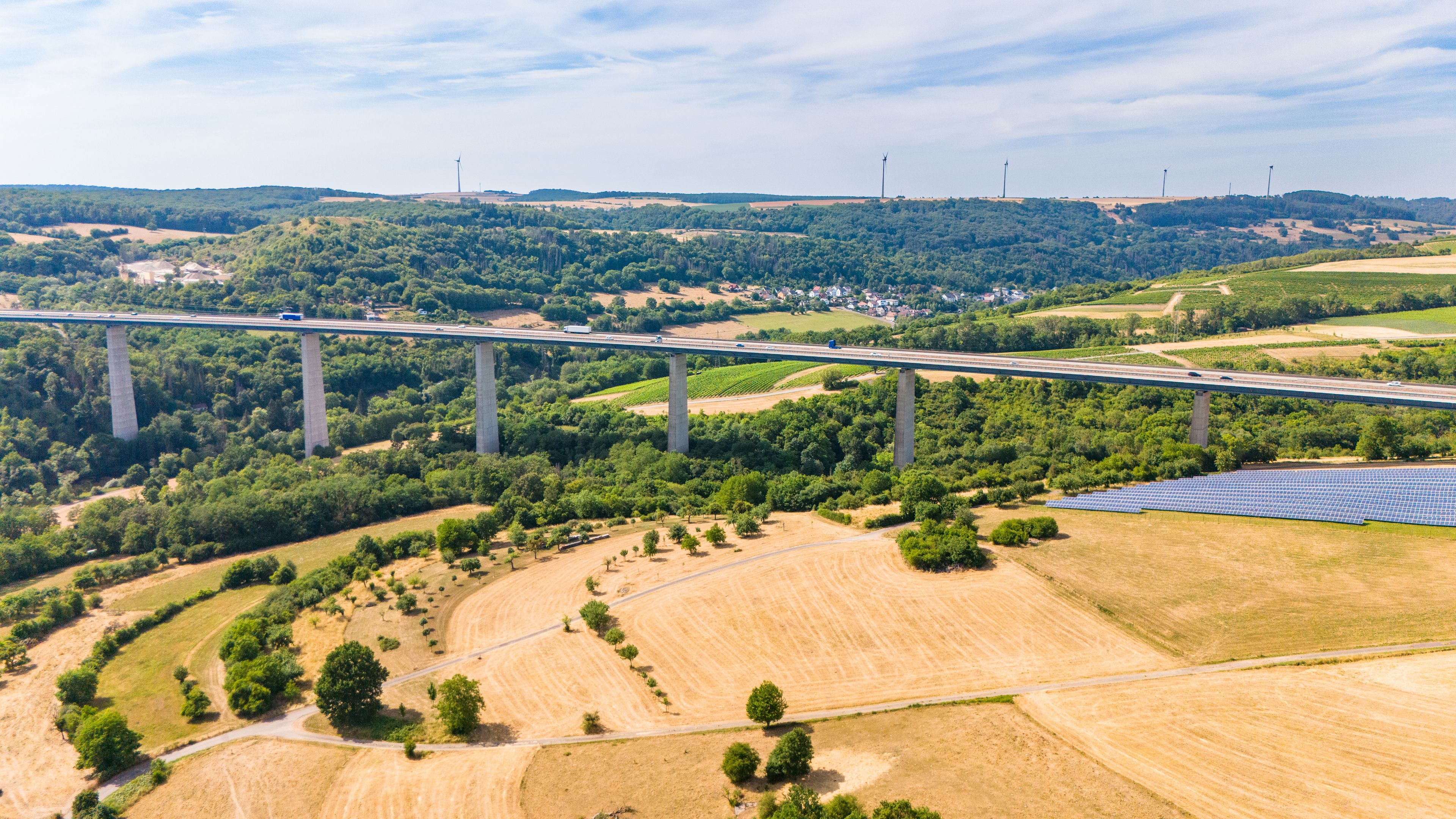 A bridge over a valley with solar panels and wind turbines in the background.