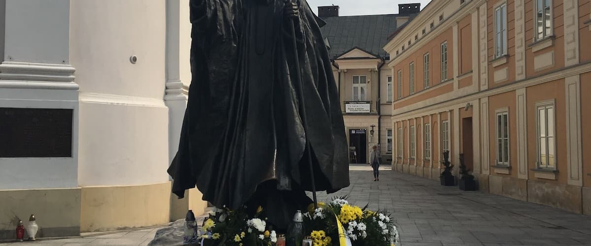 A statue of a religious figure with flowers and candles on a stone base in an urban setting.