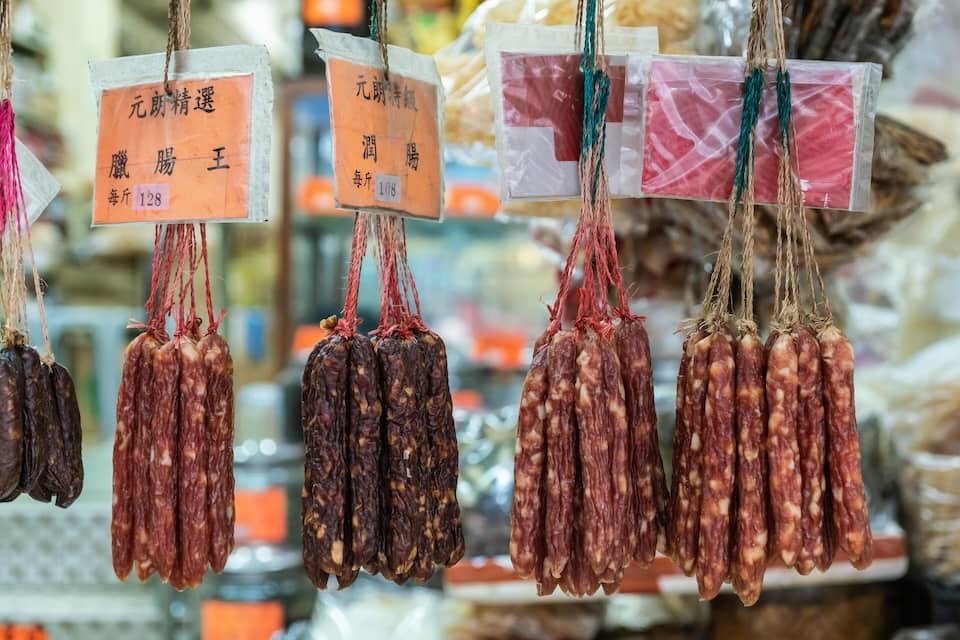 Diversi tipi di salumi secchi appesi a spago, ognuno con un'etichetta dei prezzi.