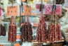 Various types of dried sausages hanging from strings, each labeled with a price tag.