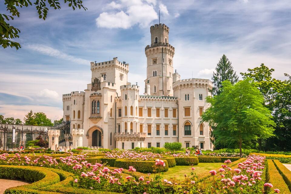 Um castelo com uma torre, rodeado por um jardim formal com flores cor-de-rosa.