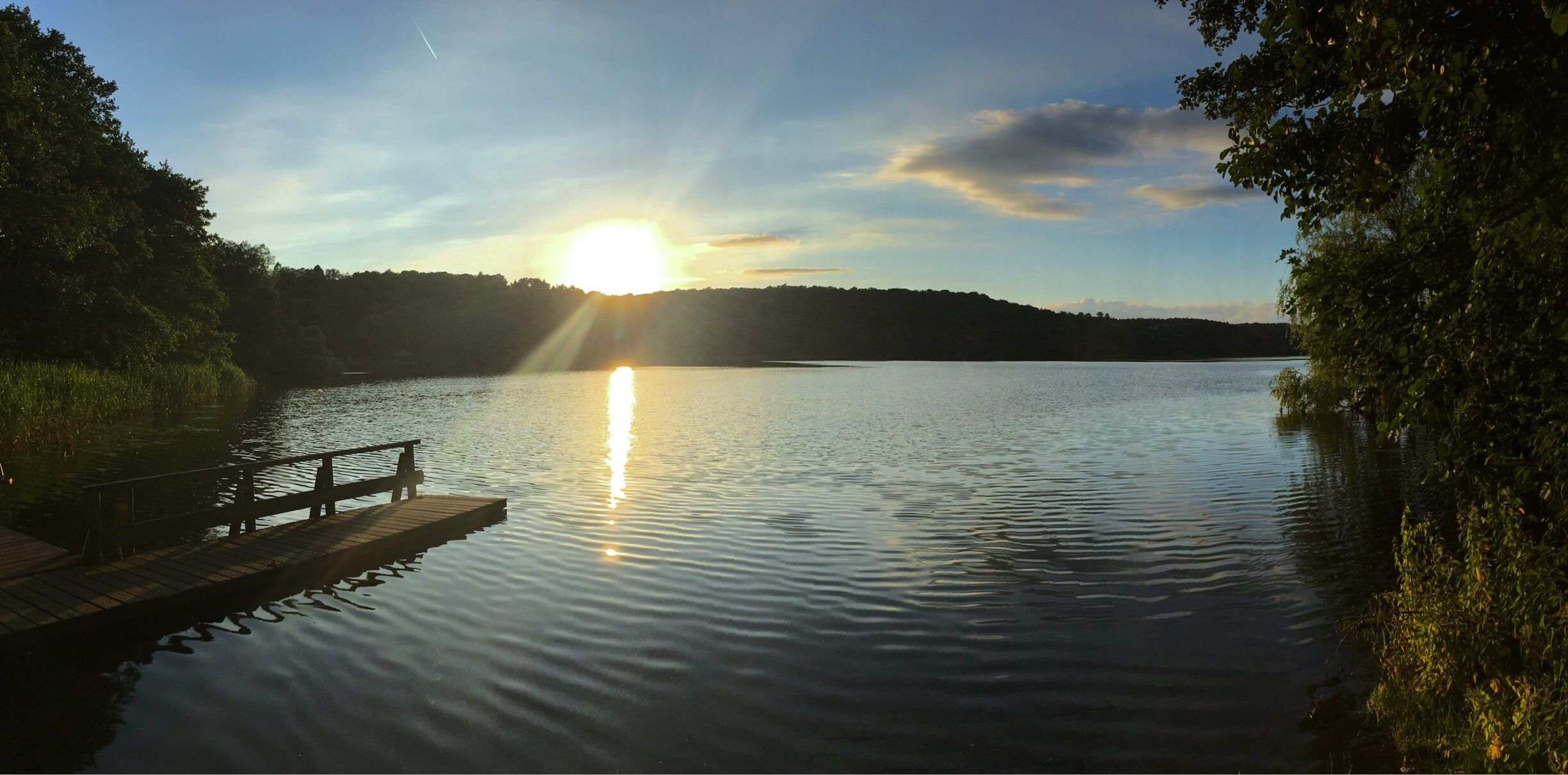A calm lake with a wooden dock extending into the water, surrounded by trees and a clear sky with the sun setting on the horizon.
