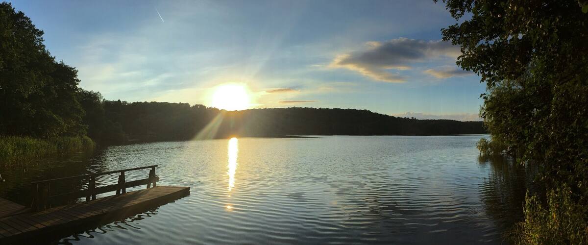 A calm lake with a wooden dock extending into the water, surrounded by trees and a clear sky with the sun setting on the horizon.