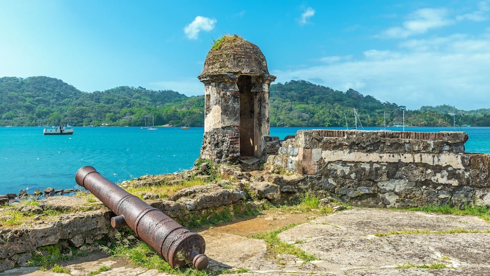A coastal fort with a cannon, a small tower, and a clear blue sea with boats in the background.