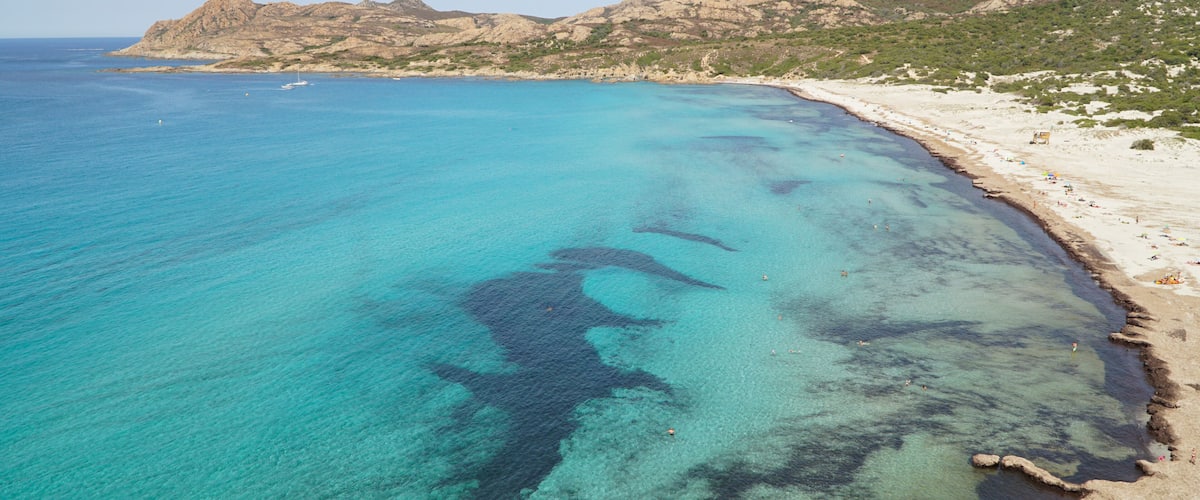 A coastal landscape with clear turquoise waters, a sandy beach, and rocky mountains in the background.