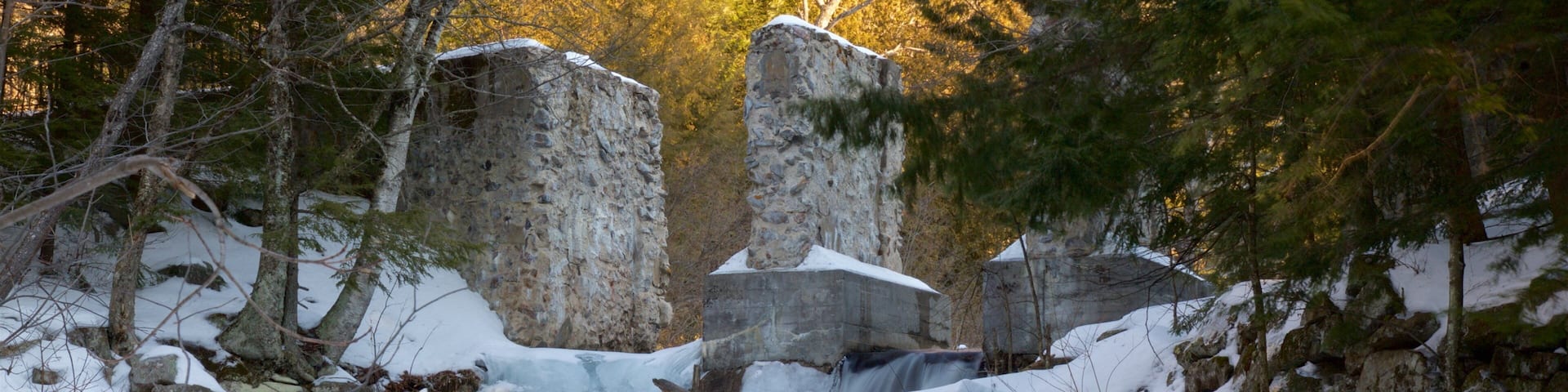 A snow-covered waterfall with large boulders and trees in the background.
