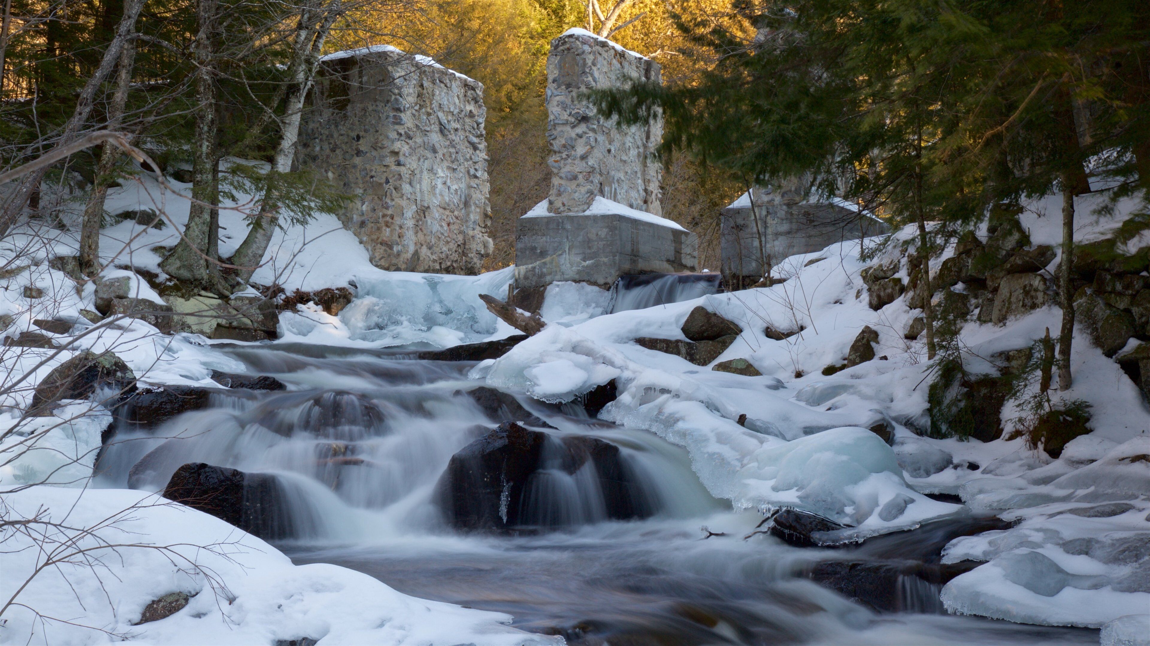 A snow-covered waterfall with large boulders and trees in the background.