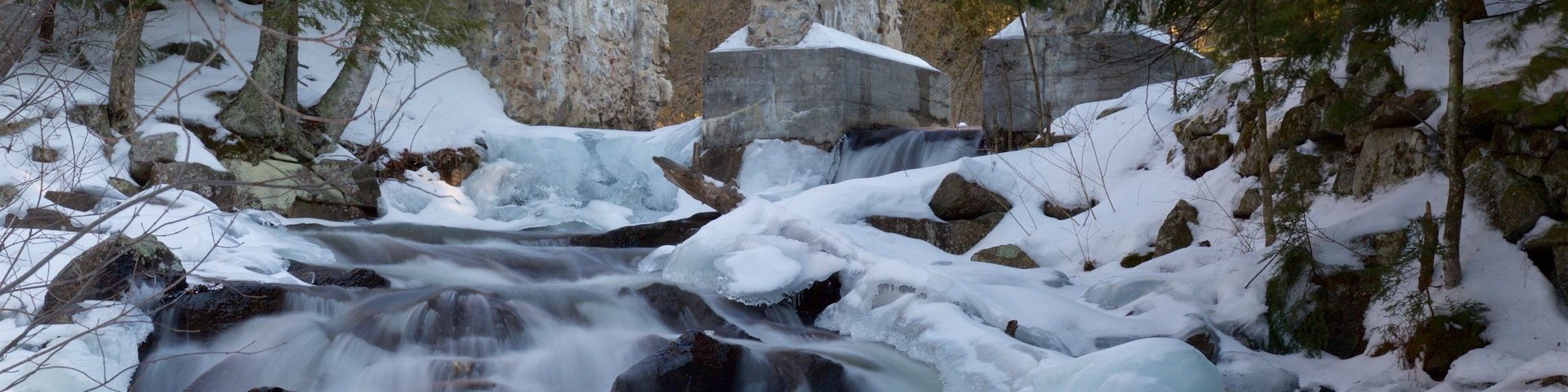 A snow-covered waterfall with large boulders and trees in the background.
