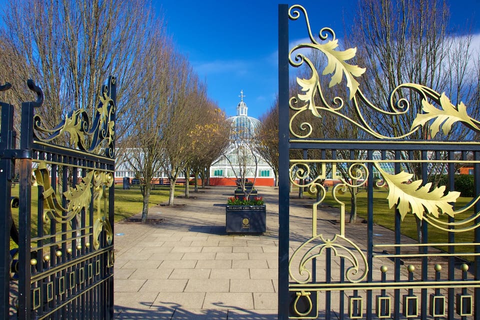 A wrought iron gate with leaf designs, a pathway leading to a building with a dome, and a clear blue sky.