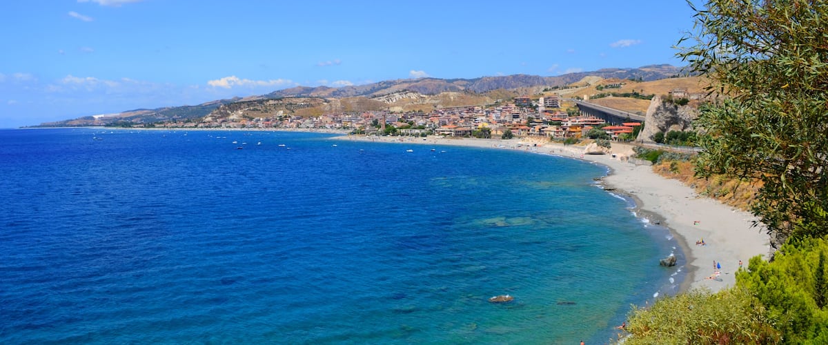 A coastal town with a beach, clear blue sea, and mountains in the background.