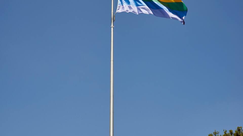 Eine Fahnenstange mit einer Regenbogenflagge vor blauem Himmel.