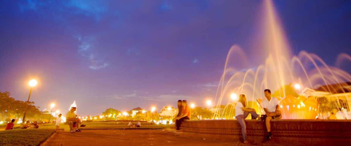 A fountain with water jets, people sitting on steps, and a park with trees and street lamps at night.