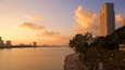 A sunset over a river with a bridge, a tall building, and a promenade with people walking.