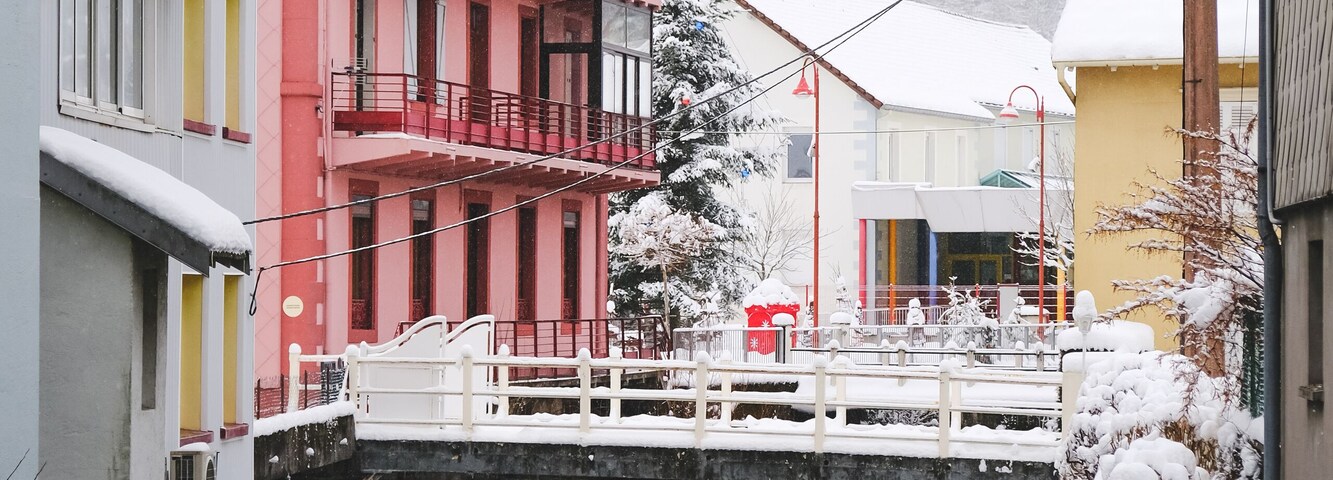 Uma ponte coberta por neve sobre um rio, edifícios coloridos e montanhas ao fundo.