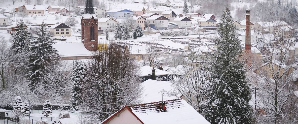 A snow-covered town with a prominent church steeple, residential buildings, and a forested mountain backdrop.