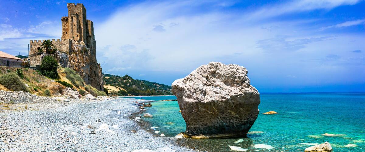 A rocky beach with a large boulder in the water, a historic stone tower, and a clear blue sky.