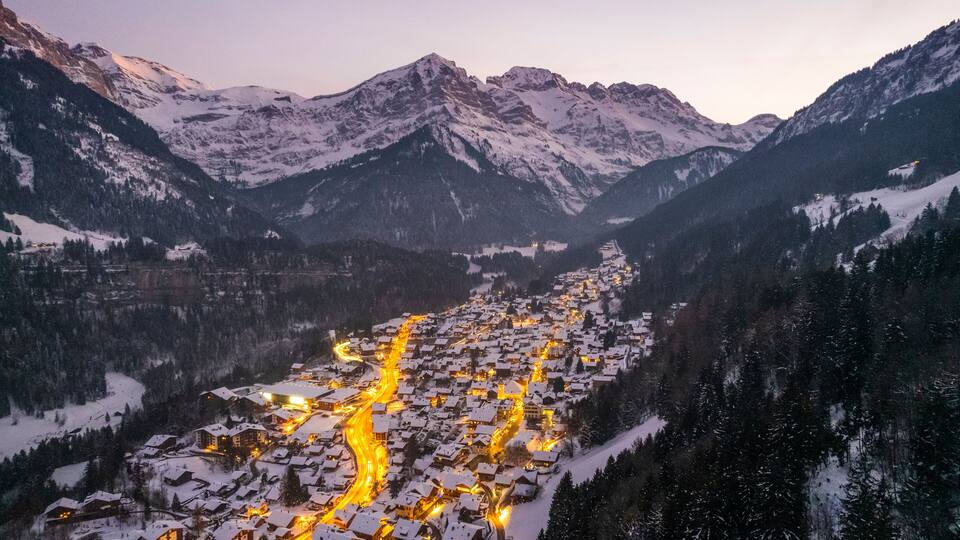 A snow-covered mountain village with winding roads and illuminated buildings at dusk.