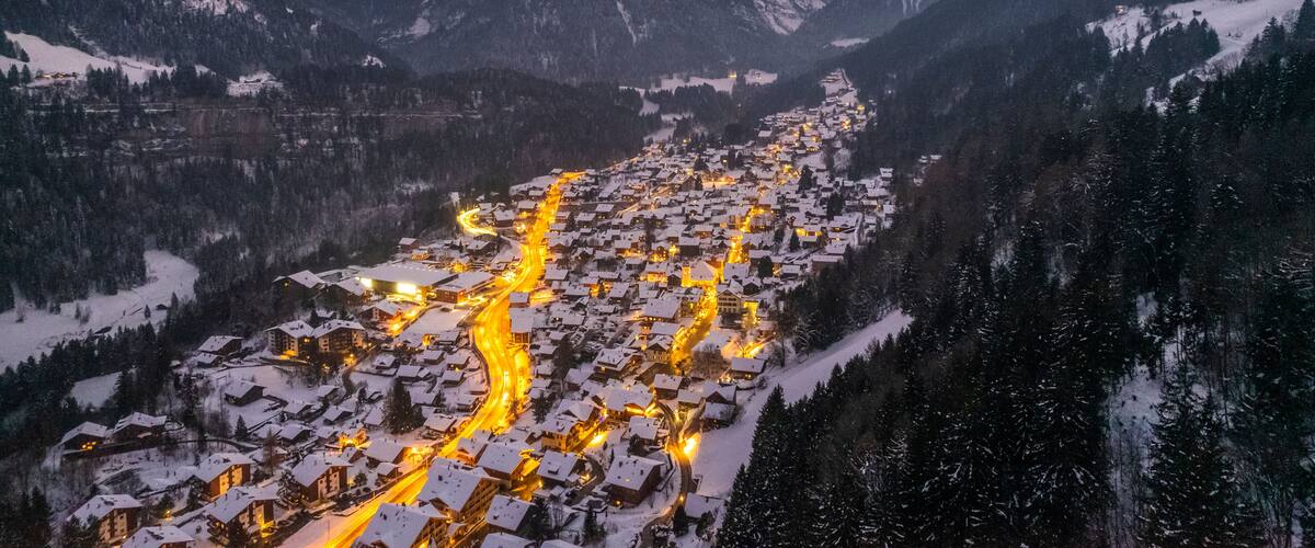 A snow-covered mountain village with winding roads and illuminated buildings at dusk.