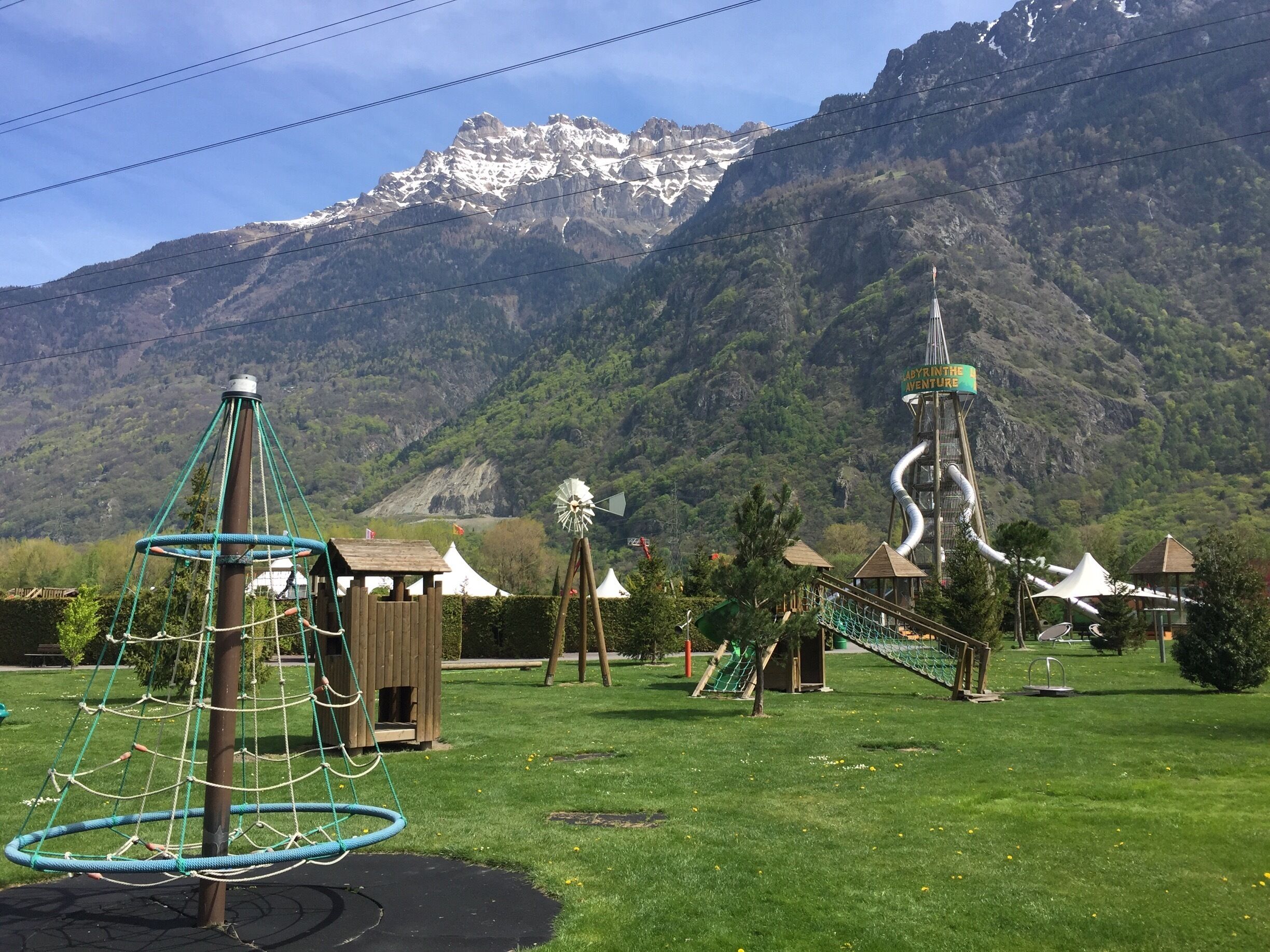 A playground with a rope swing, a climbing structure, and a windmill in a park with a mountain backdrop.