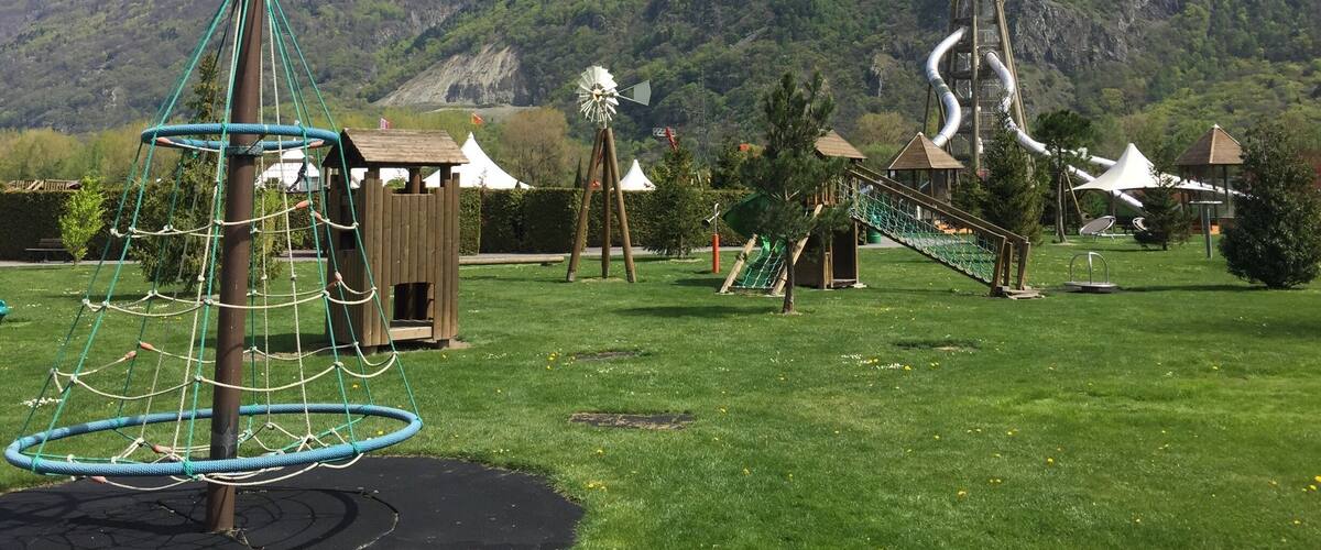 A playground with a rope swing, a climbing structure, and a windmill in a park with a mountain backdrop.