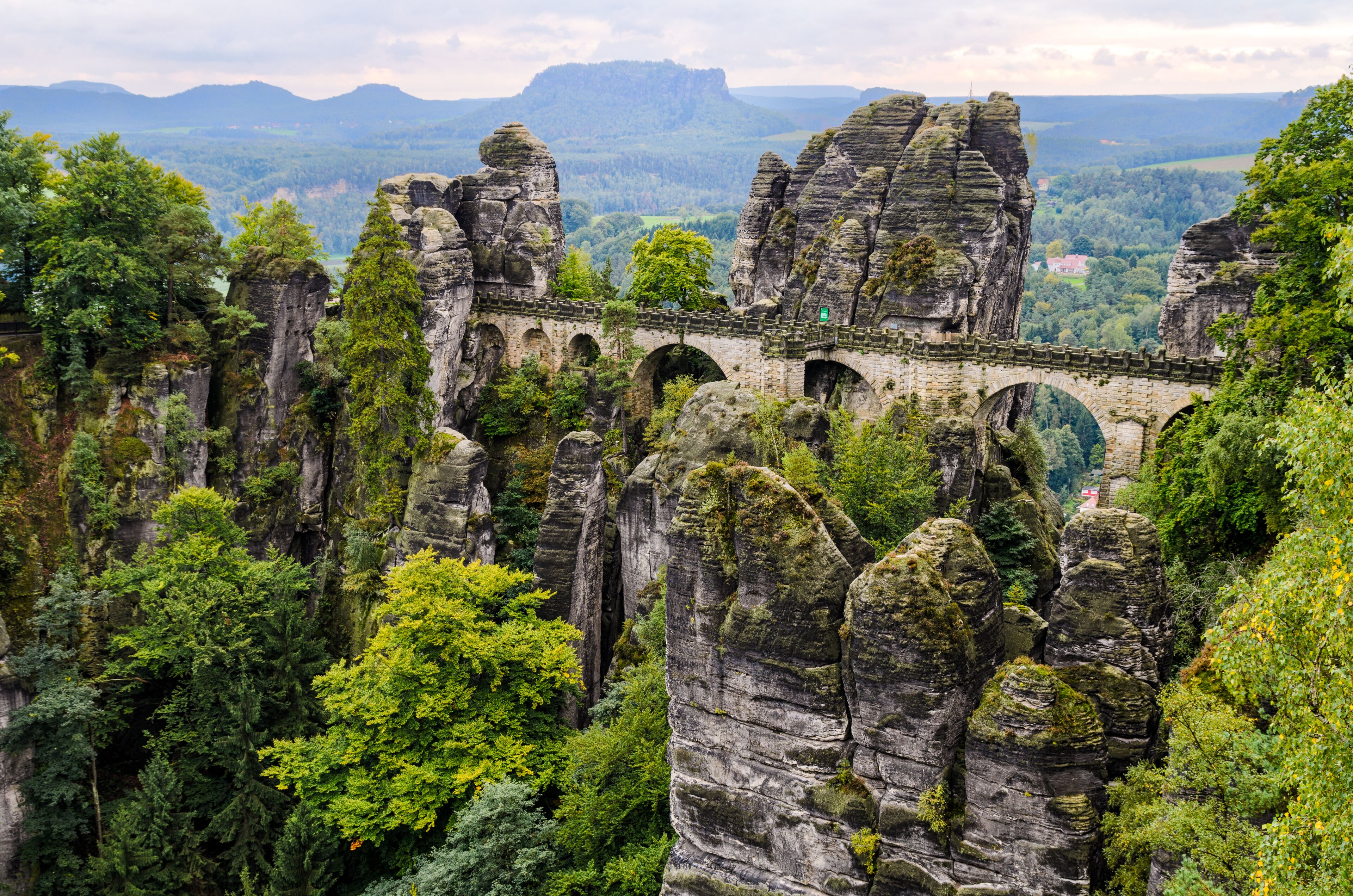 A stone bridge over a gorge with towering rock formations and dense green forest.