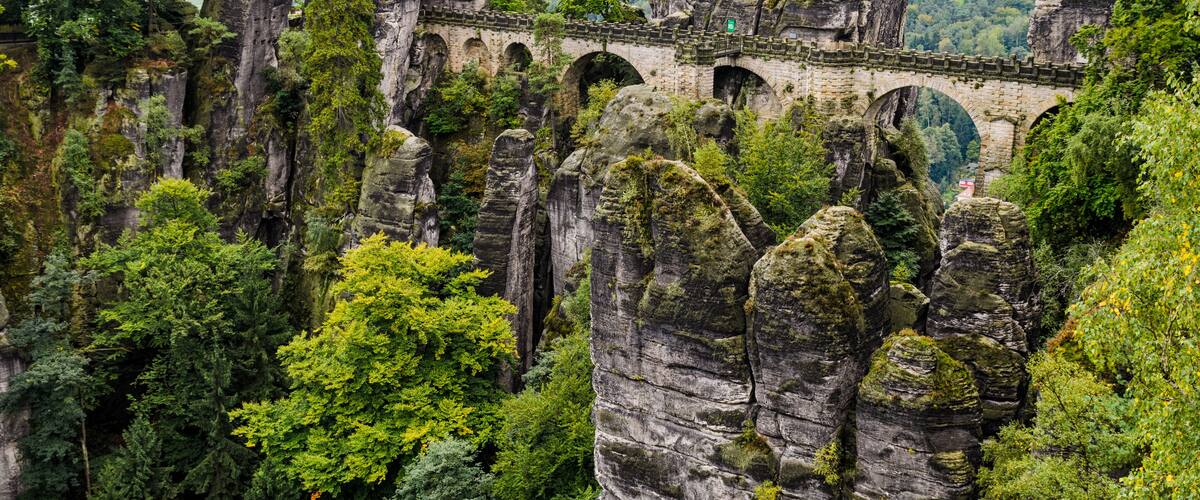 A stone bridge over a gorge with towering rock formations and dense green forest.