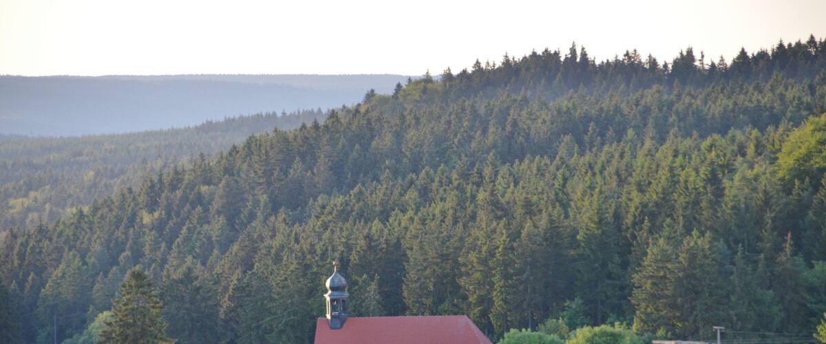 A white church with a red roof surrounded by a forested hillside.