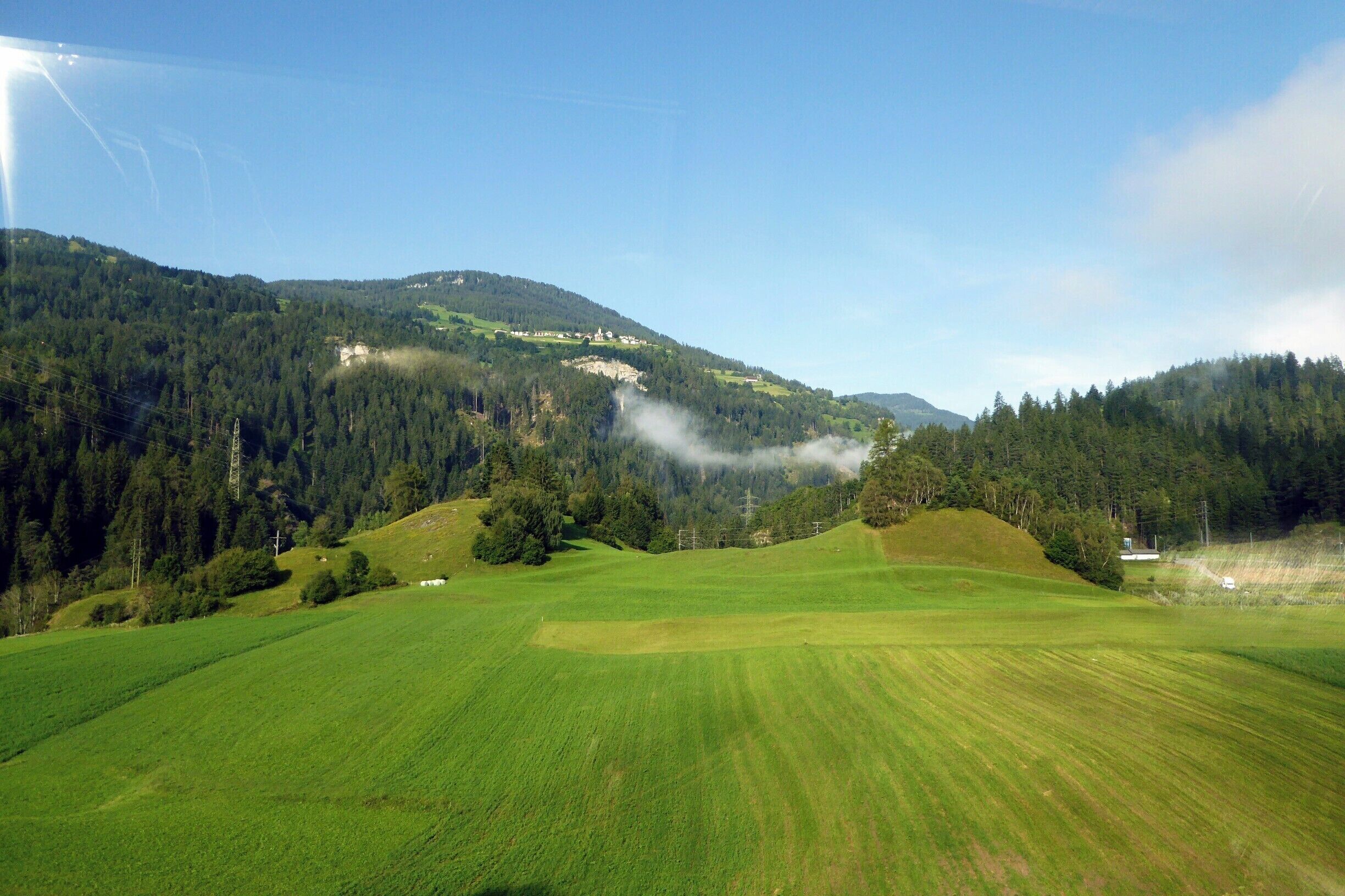 A green valley with rolling hills, a forested mountain, and a small building in the distance.
