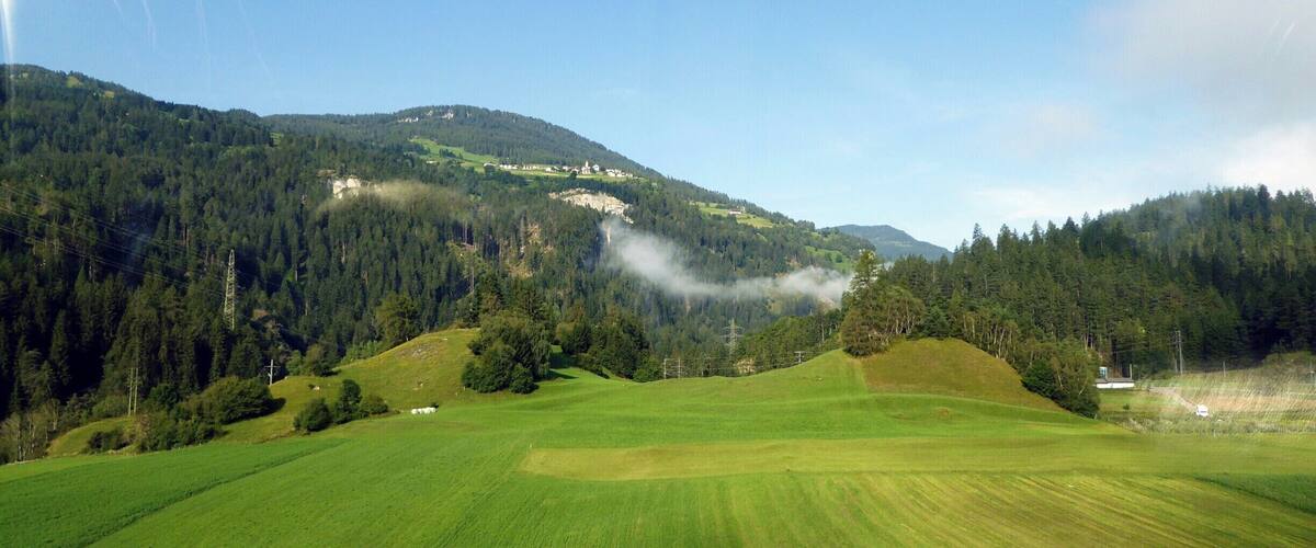 A green valley with rolling hills, a forested mountain, and a small building in the distance.