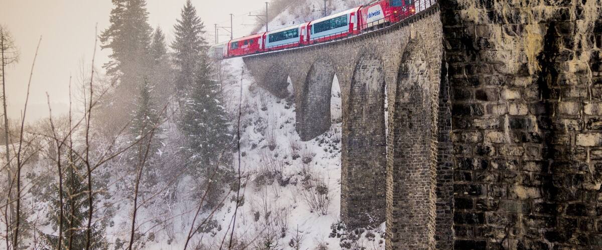 A red and white train on a stone bridge in a snowy forest.