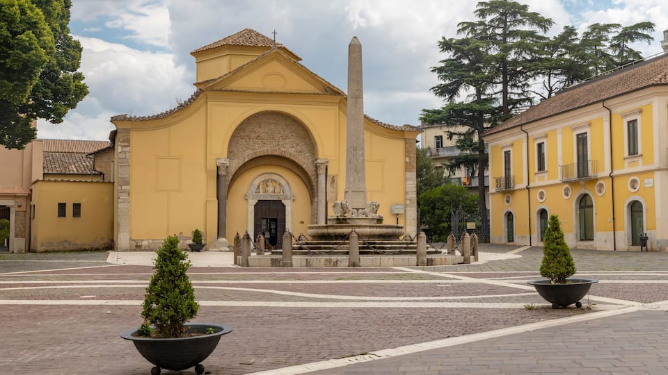 A historic yellow building with an obelisk, a fountain, and two potted trees in a paved square.