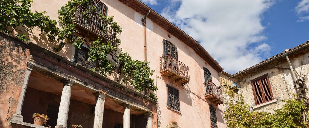 A historic building with a pink facade, a balcony with plants, and a staircase leading to the entrance.