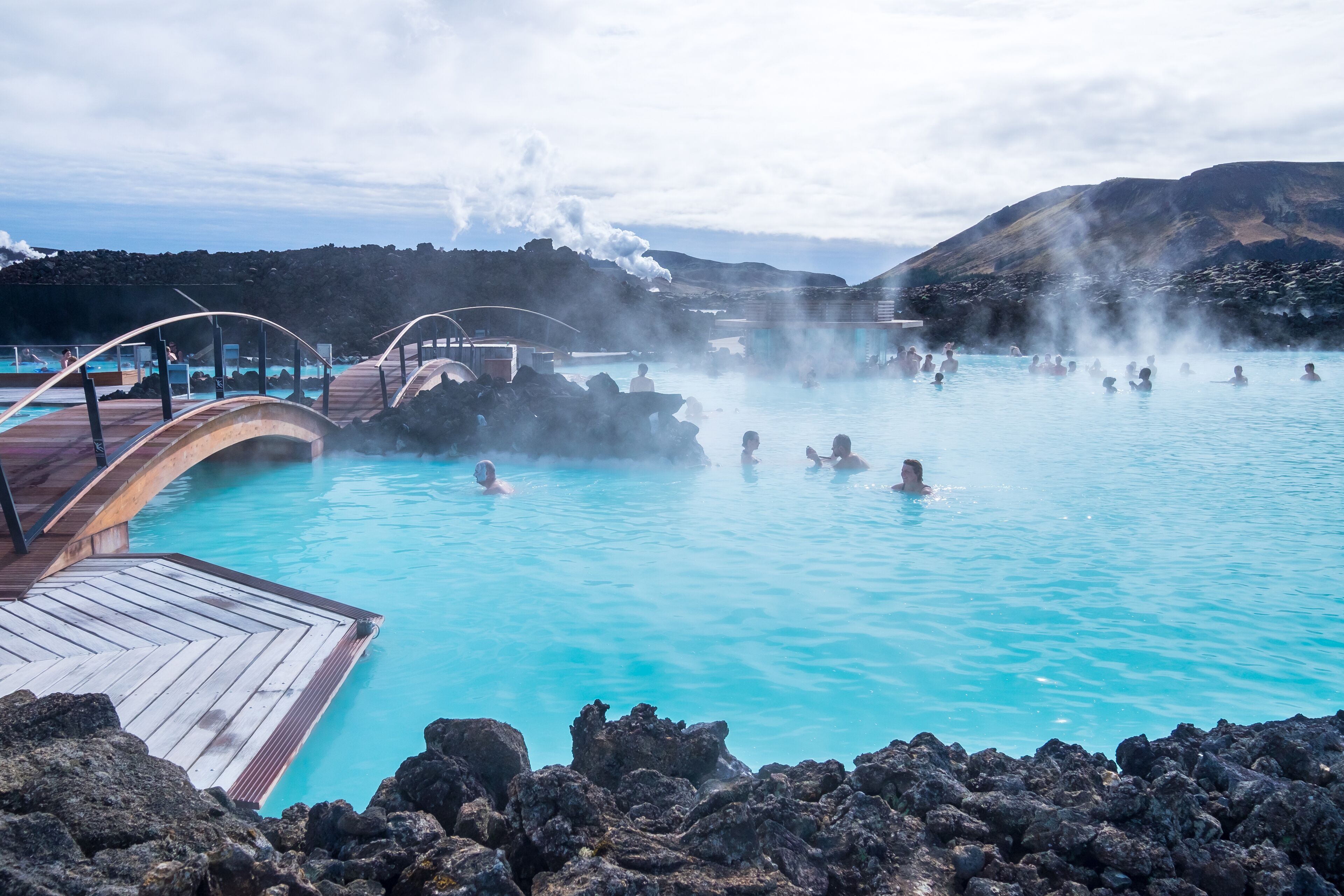 Un spa geotérmico con gente en el agua, vapor ascendiendo del suelo y una terraza de madera con vistas a la piscina.