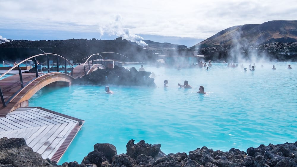 A geothermal spa with people in the water, steam rising from the ground, and a wooden deck overlooking the pool.