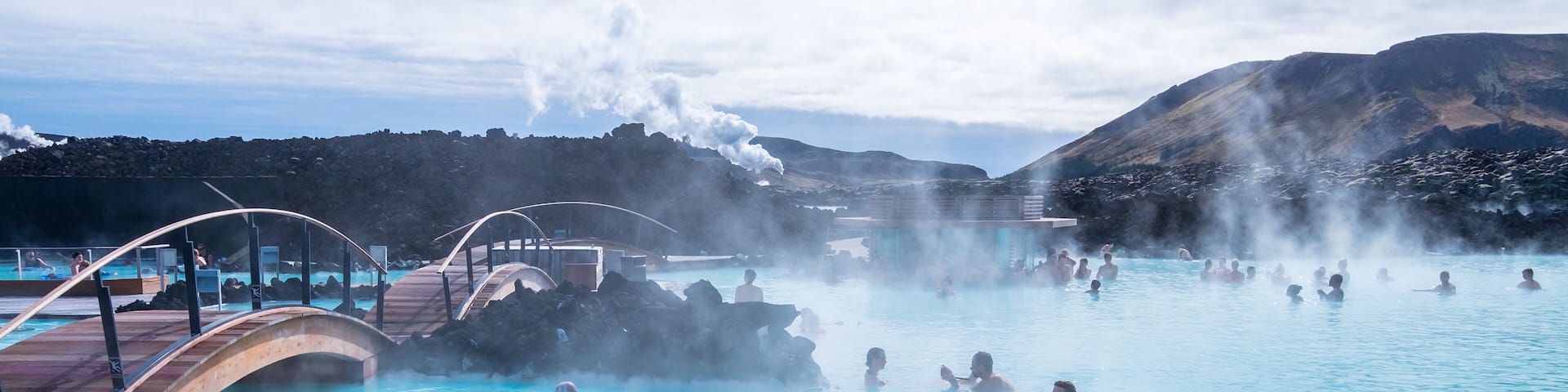 A geothermal spa with people in the water, steam rising from the ground, and a wooden deck overlooking the pool.