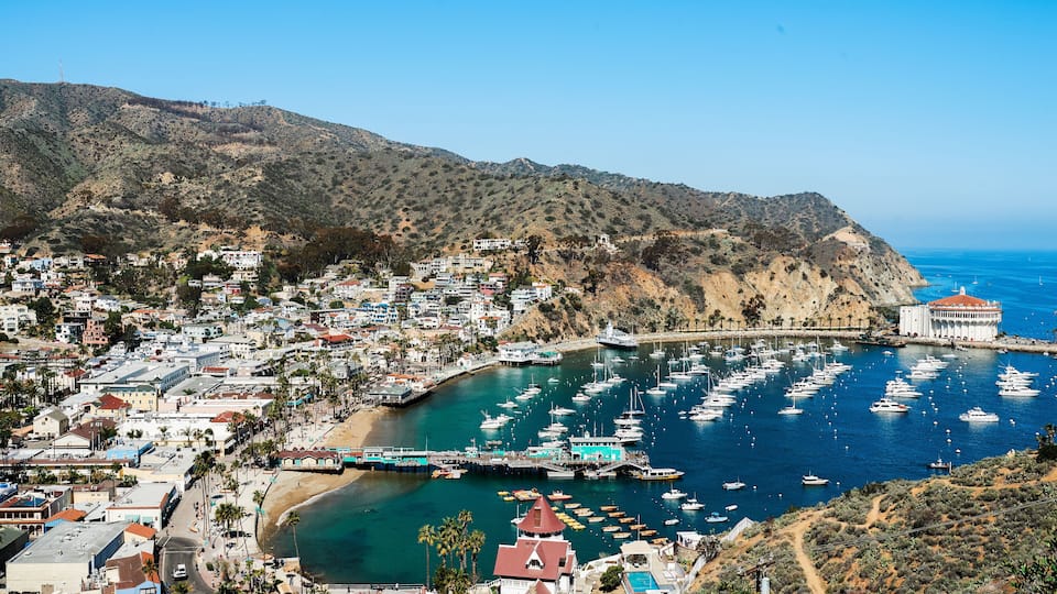 A coastal town with a marina filled with boats, a beach, and a mountainous backdrop.