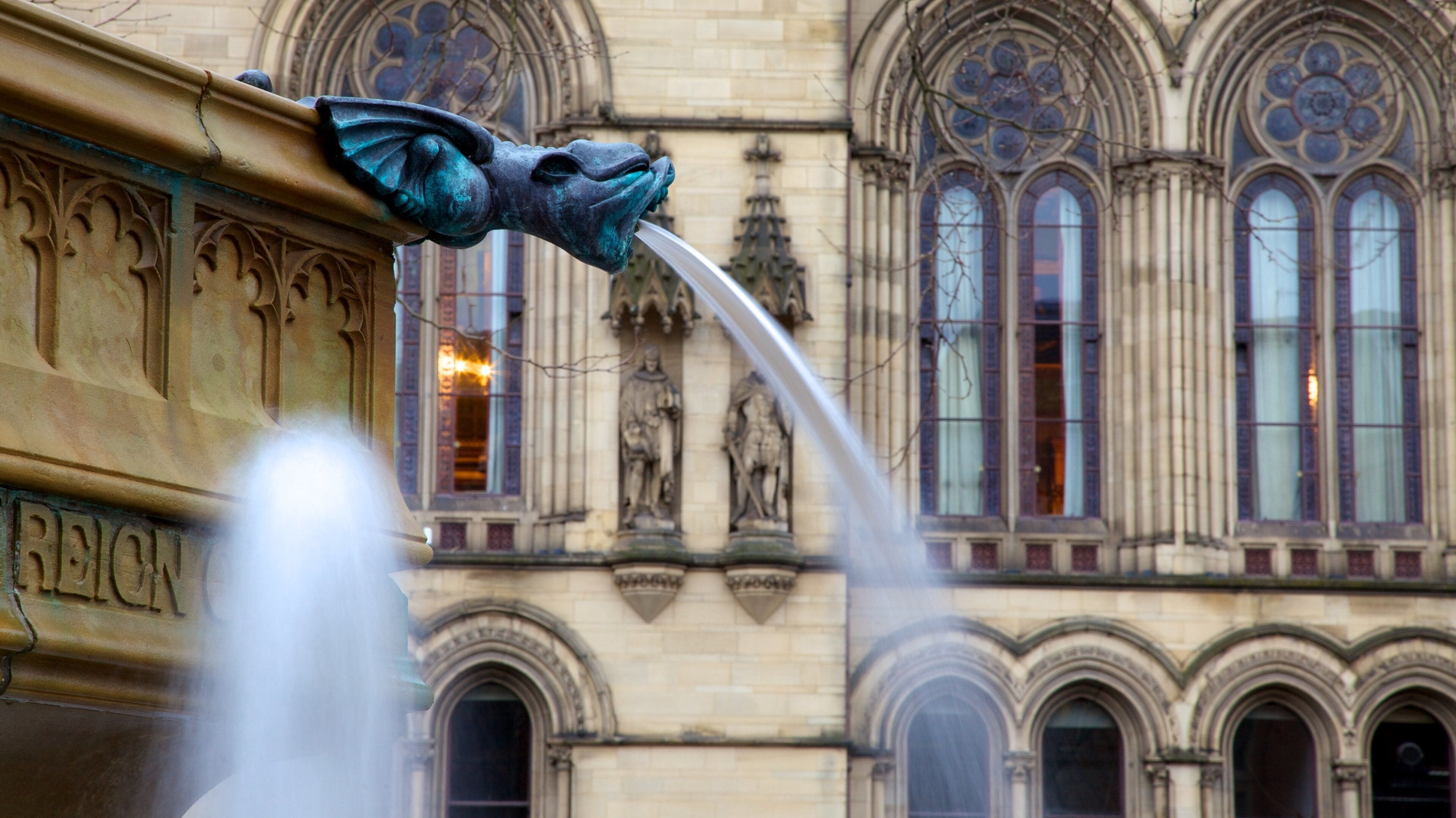 A bronze water spout with a lion's head, a building with arched windows, and a statue in the background.