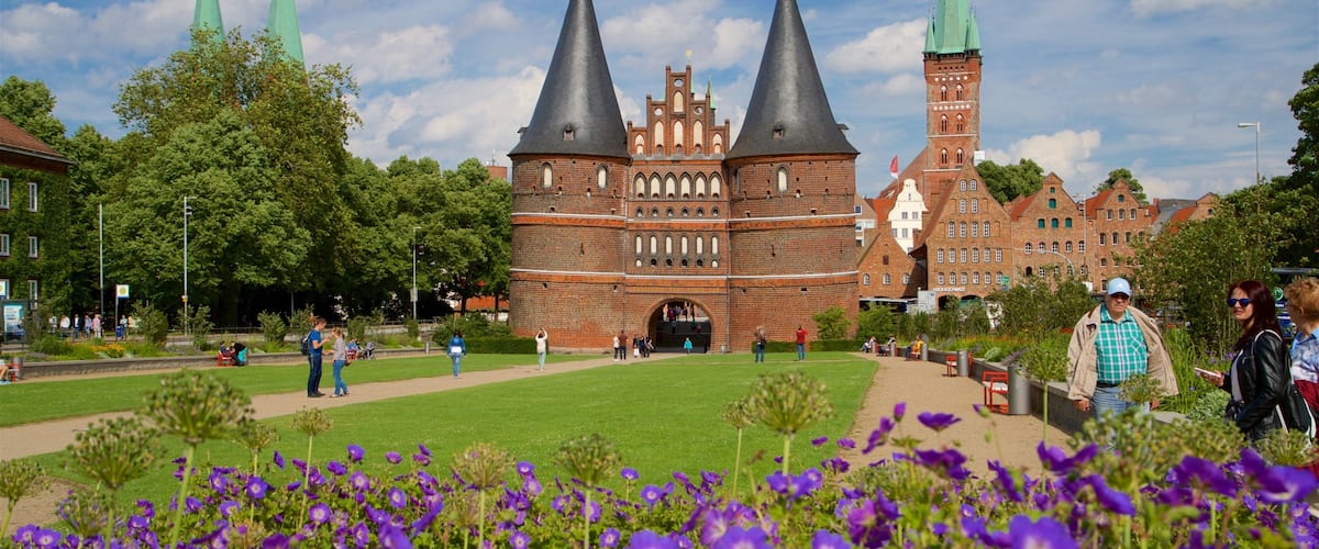 A historic building with two tall spires and a brick tower, surrounded by a park with purple flowers.