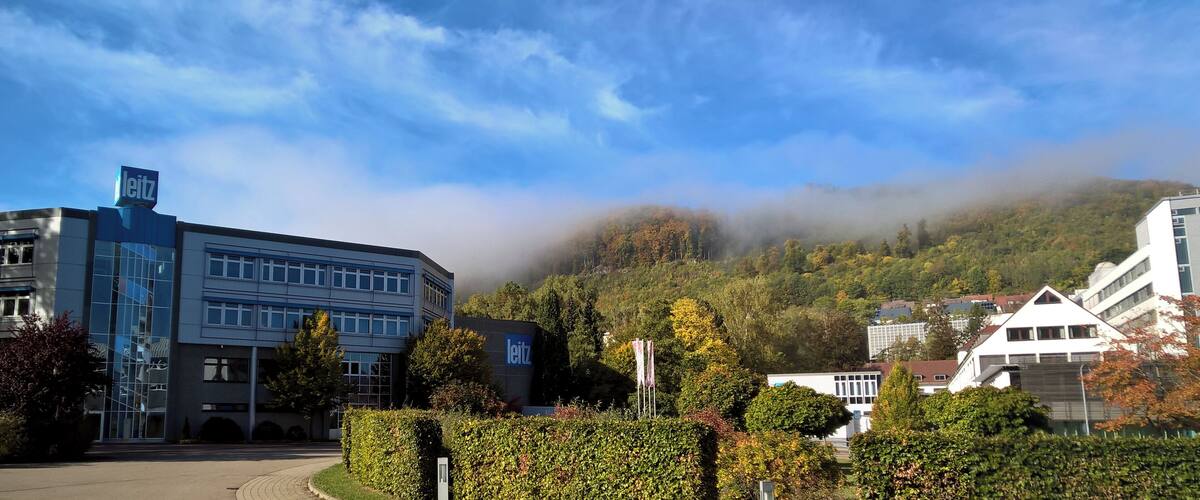 A modern building with a 'Leitz' sign, surrounded by lush greenery and a forested hill in the background.