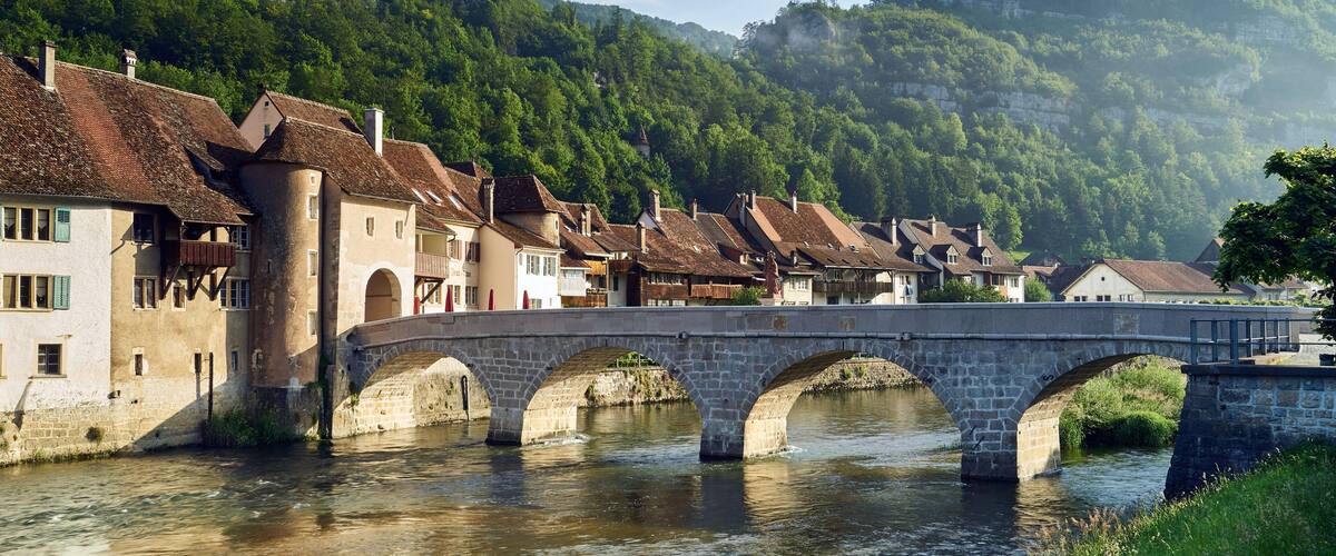 A stone bridge over a river, surrounded by traditional buildings with sloped roofs, set against a backdrop of lush green hills.