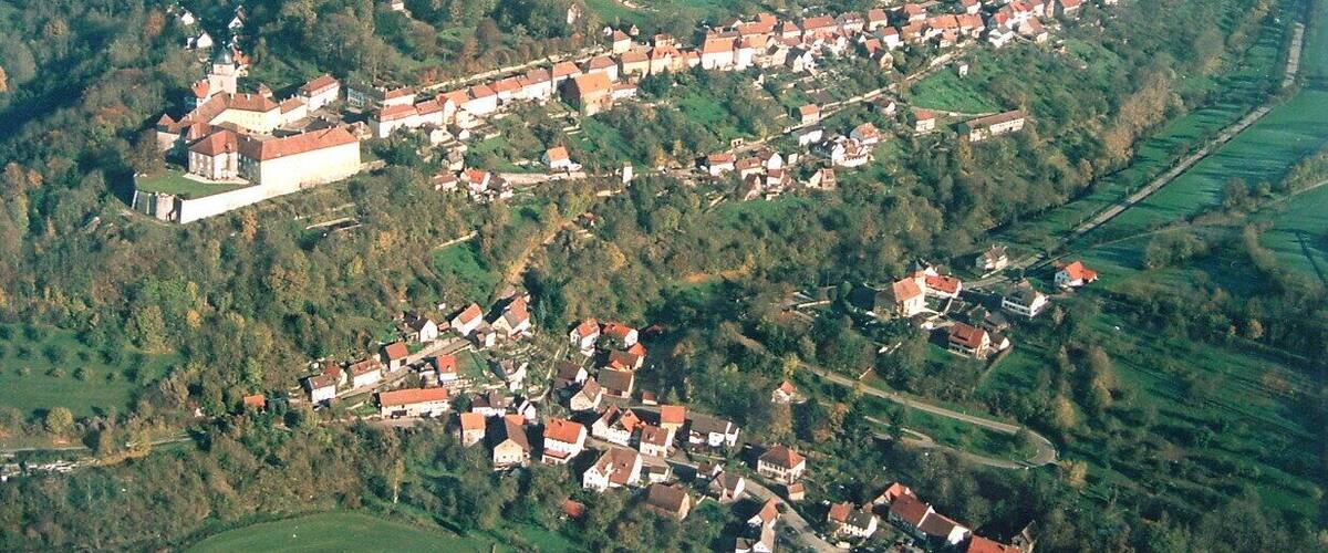 An aerial view of a village with a prominent castle on a hill, surrounded by green fields and scattered houses.