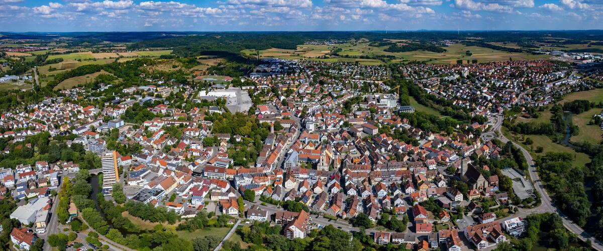 An aerial view of a densely populated town with a mix of residential and commercial buildings, surrounded by green fields and distant wind turbines.