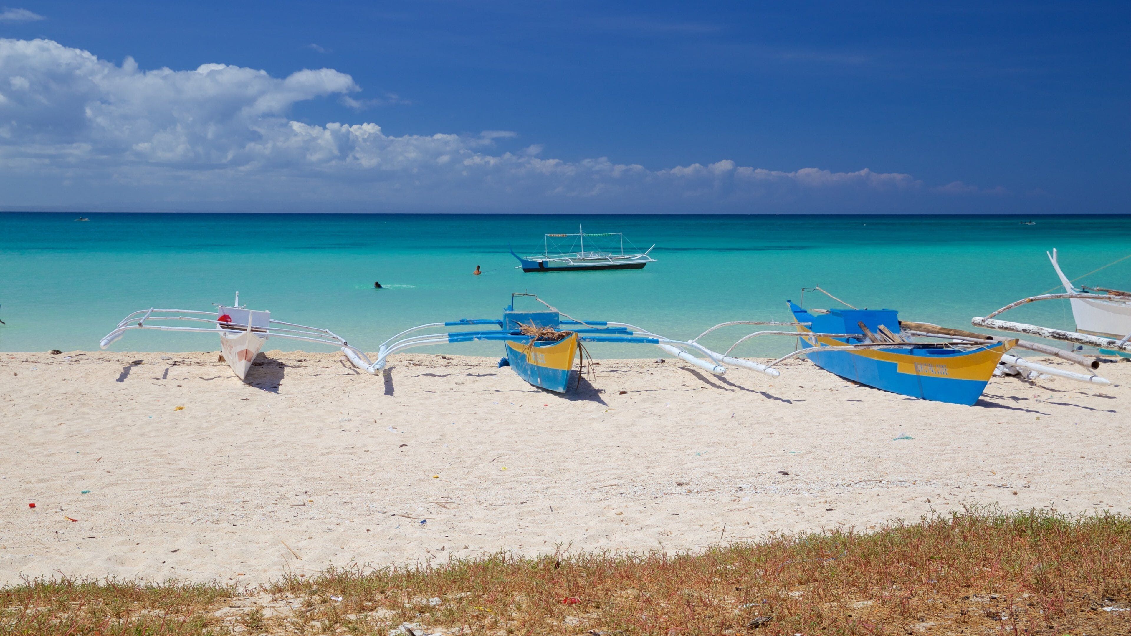 A beach with several boats, clear turquoise water, and a few people swimming.