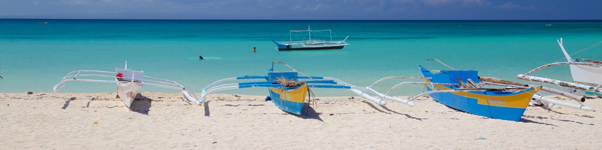 Uma praia com vários barcos, água turquesa transparente e algumas pessoas nadando.