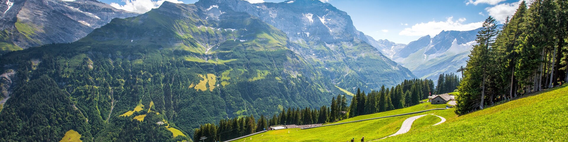 Paisagem montanhosa com estrada sinuosa, campos verdes e um pequeno edifício.