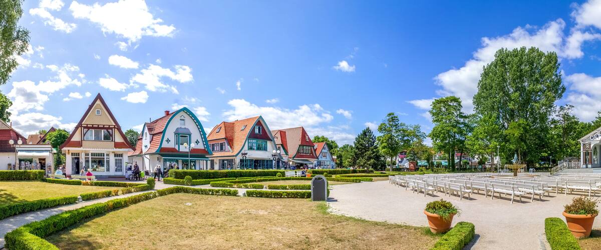 A park with neatly trimmed hedges, a large open space with rows of white benches, and traditional buildings with red roofs.